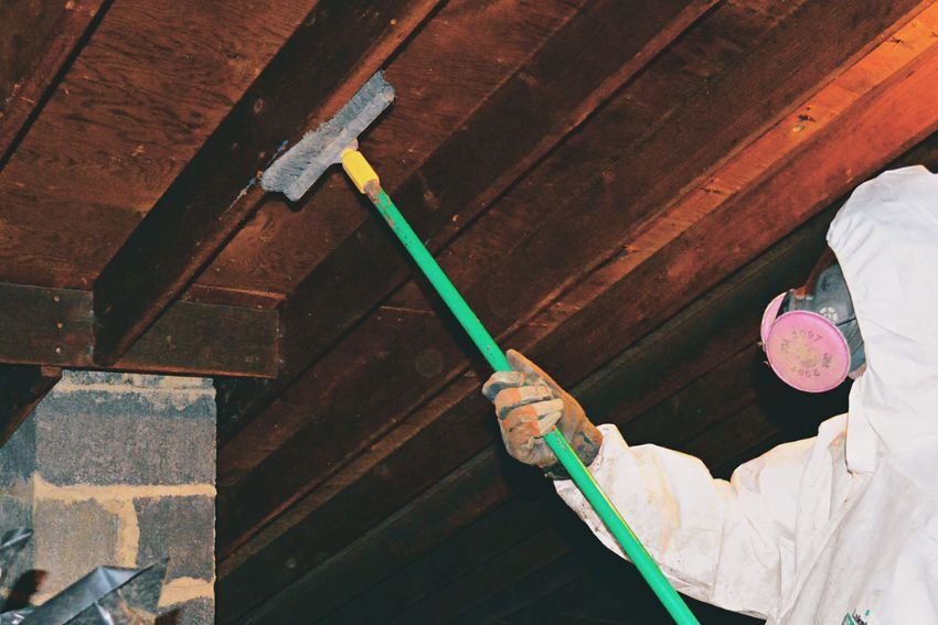 A man in a white protective suit is seen cleaning a wooden ceiling, representing attic mold removal in Las Vegas, NV.